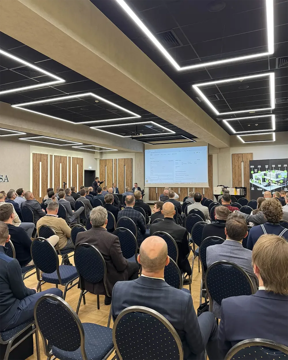 Seated audience in a conference hall with linear ceiling lights, watching a presentation projected on a screen at KNX Experts Meeting 2024