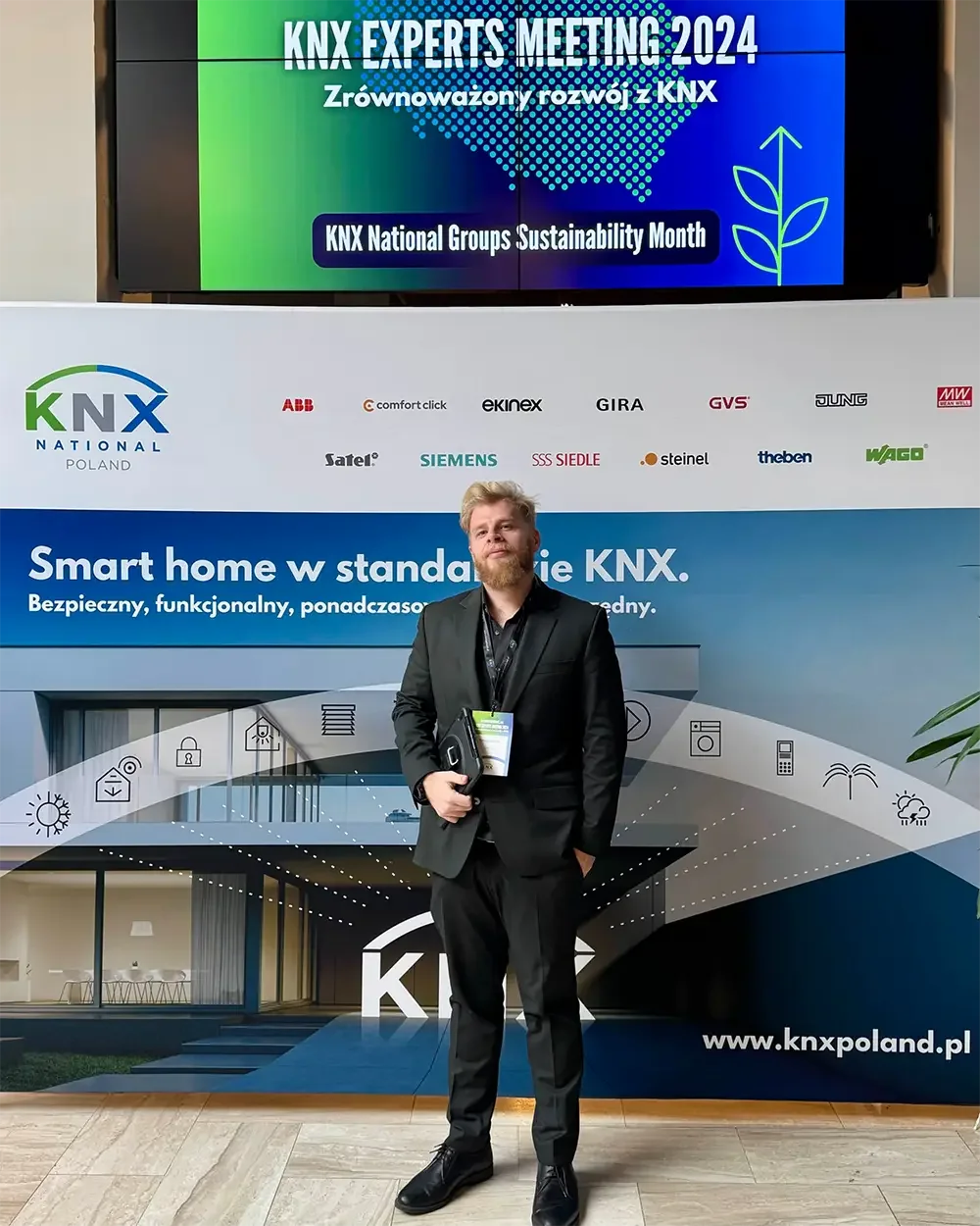 Man in a black suit with a delegate badge, holding a tablet, in front of the KNX National Poland partner wall, under a screen reading KNX Experts Meeting 2024