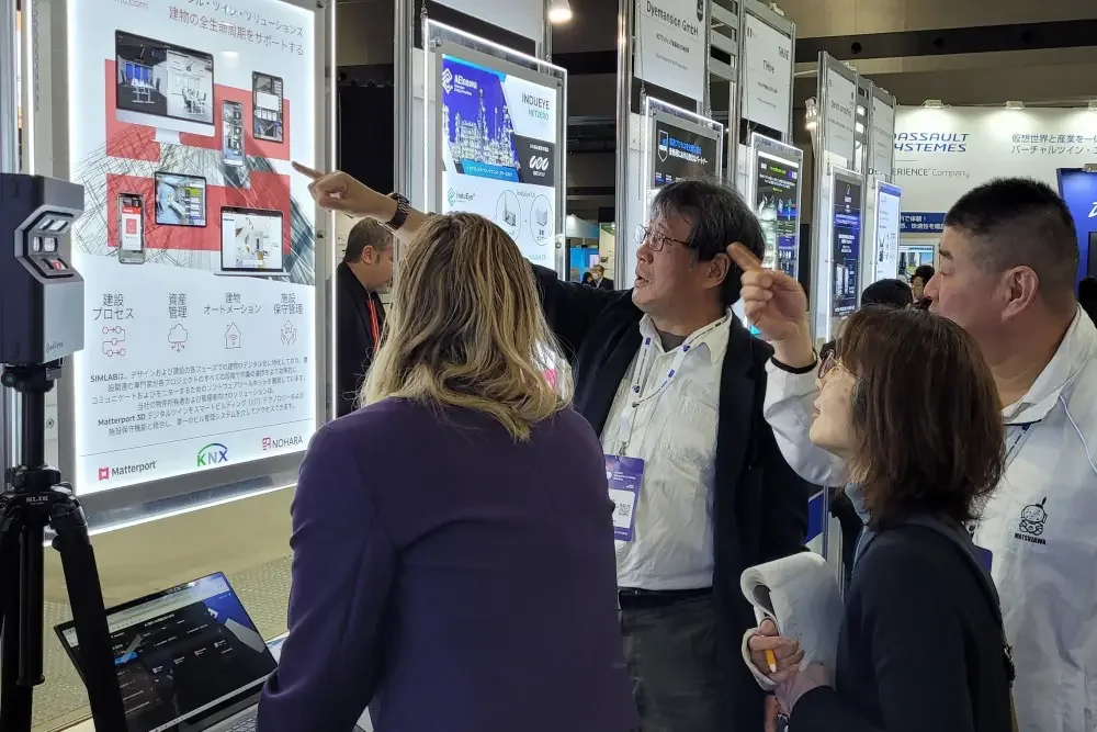 Visitors at the EU-Japan Centre booth looking at a Japanese-language SIMLAB poster as a man points at it; a 3D camera on a tripod in front
