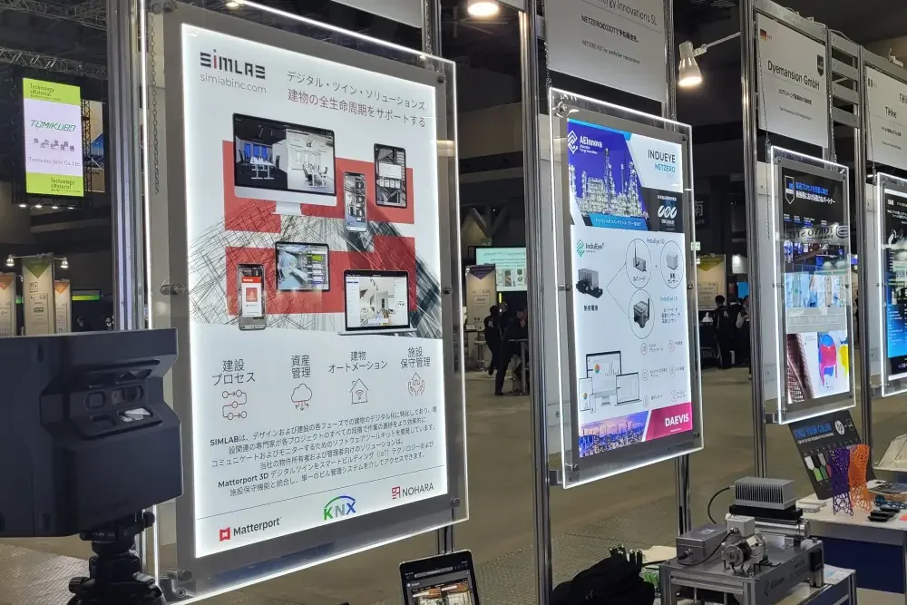 Illuminated Japanese-language SIMLAB poster with Matterport, KNX and NOHARA logos beside other exhibitor posters; a 3D camera in the foreground