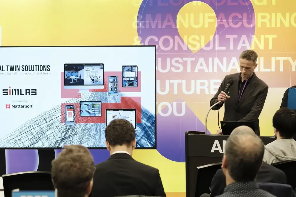 Speaker with a microphone at a podium next to a screen showing a SIMLAB Digital Twin Solutions slide powered by Matterport, audience in front