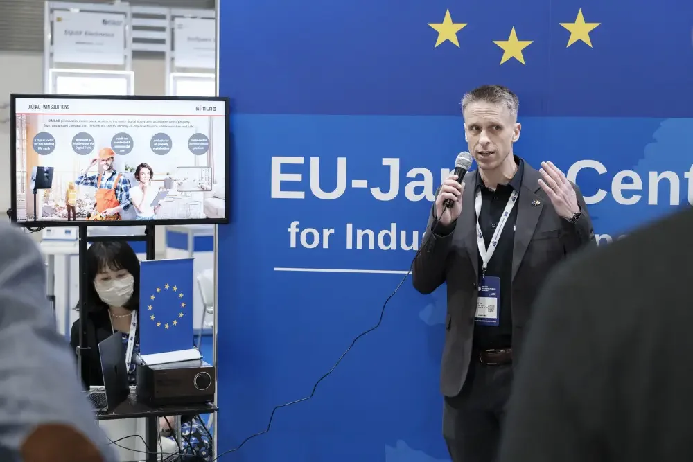 Speaker with a microphone in front of the EU-Japan Centre for Industrial Cooperation backdrop, beside a screen with a SIMLAB Digital Twin Solutions slide