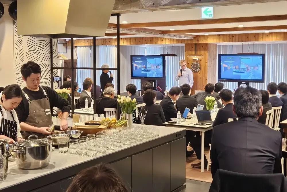 Seminar in a kitchen studio: a presenter speaks in front of two screens showing a 3D interior view while cooks prepare food at the counter