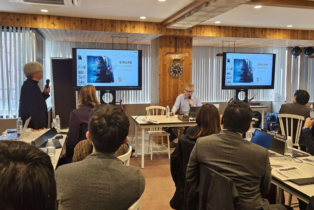 Seminar in a wood-panelled room: a seated presenter speaks into a microphone, another person stands holding one, two screens show a SIMLAB slide, audience at tables