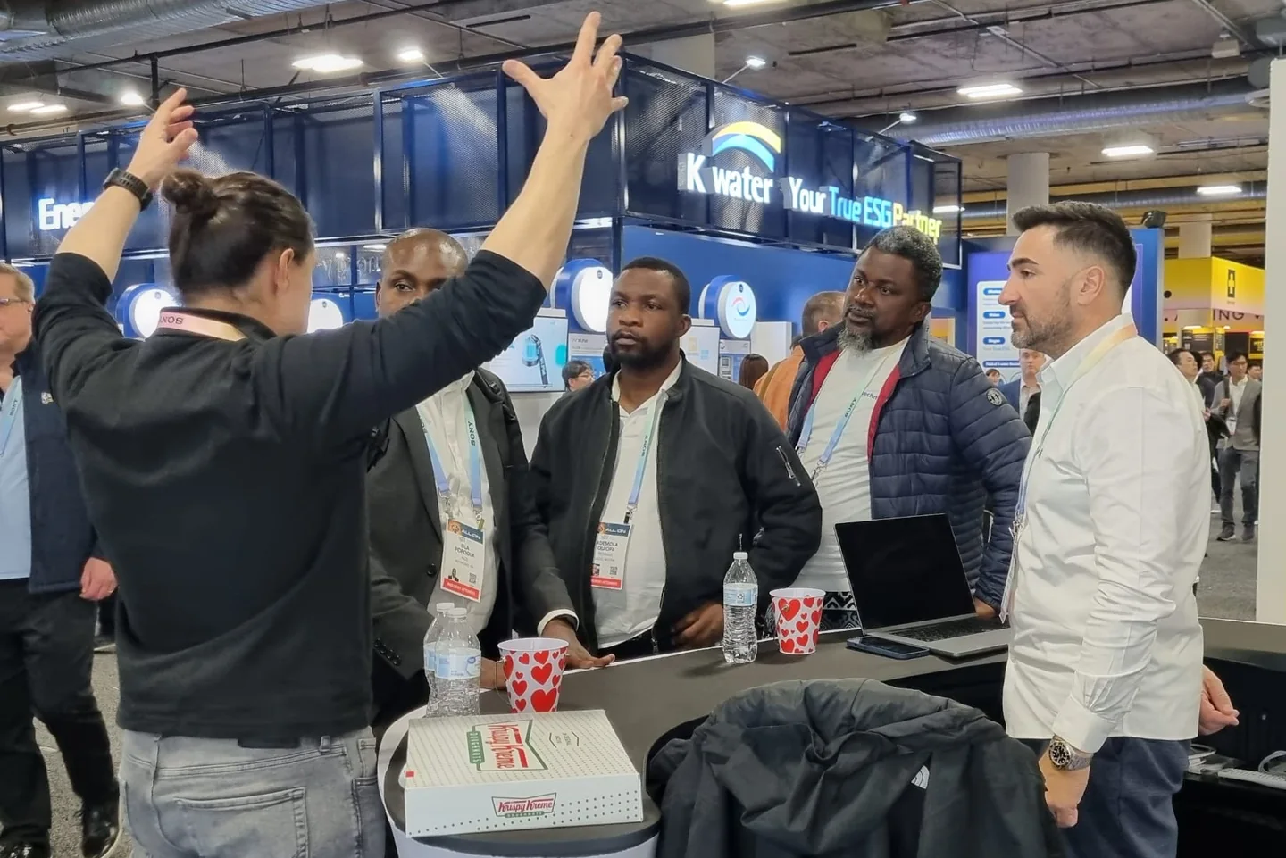 Exhibitor with raised arms explaining something to a group of visitors at a booth table at CES, the Kwater booth in the background