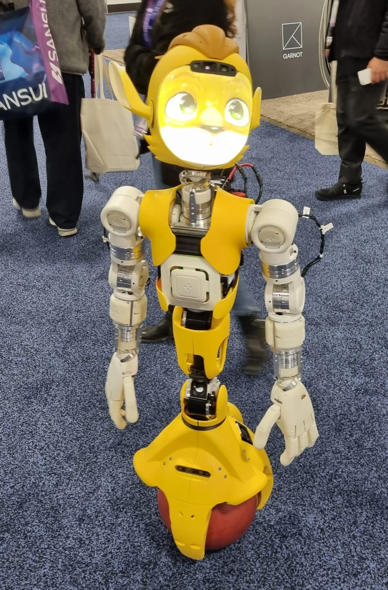 Yellow humanoid robot with an animated cat-like face, balancing on a red ball on the CES show floor