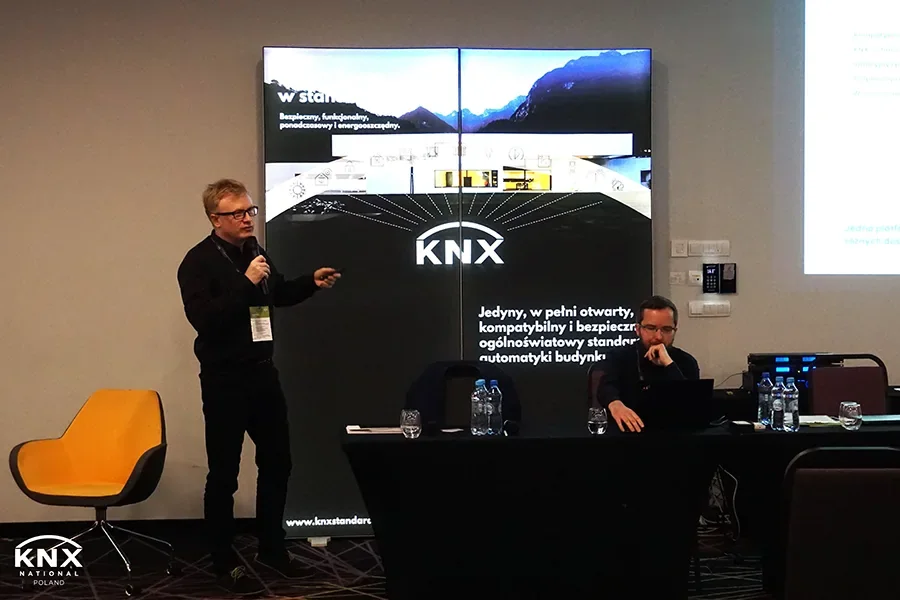 SIMLAB CEO Marek Kozlak speaking with a microphone in front of a KNX display wall at KNX Experts Meeting 2023, with a second man seated at the presenters' table with a laptop