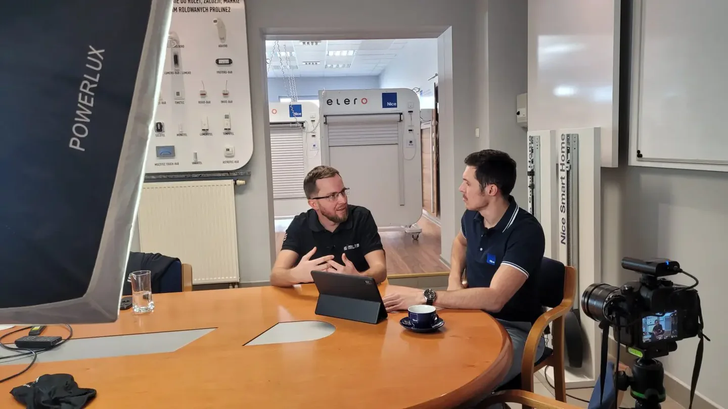A SIMLAB team member and a Nice representative talking at a table with a tablet while being filmed, in a Nice showroom with roller blind displays behind them