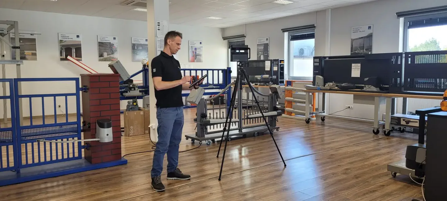 SIMLAB team member using a tablet beside a Matterport camera on a tripod while scanning a Nice training room with gate and barrier automation demo stands