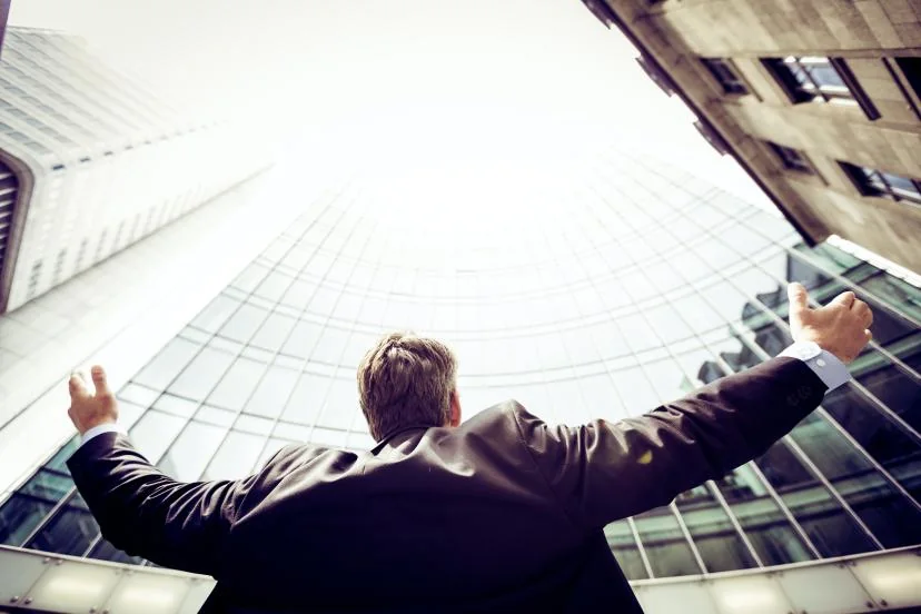 Businessman in a suit seen from behind with arms raised in celebration, looking up at glass office towers