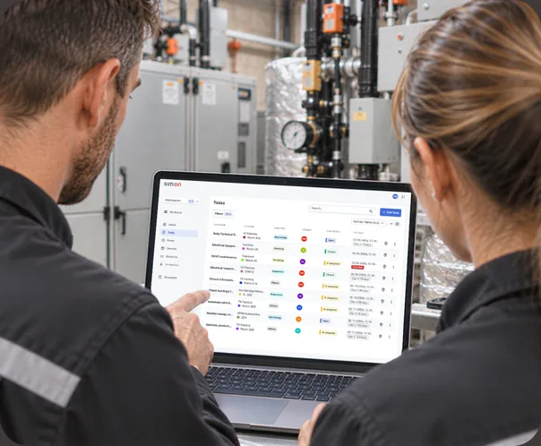 Two facility staff seen from behind in a plant room, one pointing at a SIM-ON task list on a laptop.