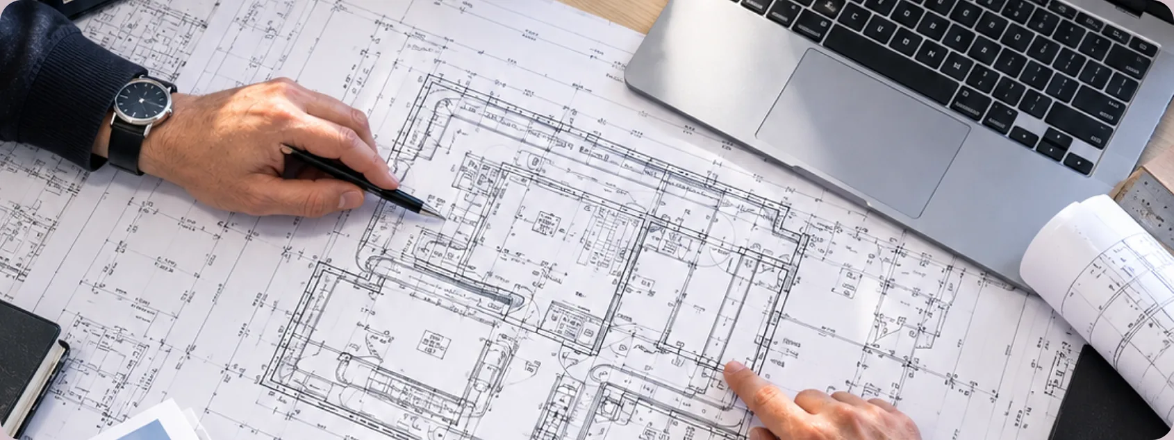 Hands marking up a printed building floor plan with a pen beside a laptop and rolled drawings