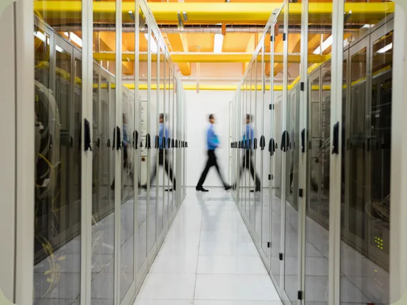 Data center aisle between glass-fronted server cabinets under yellow cable trays, with a technician walking past in motion blur